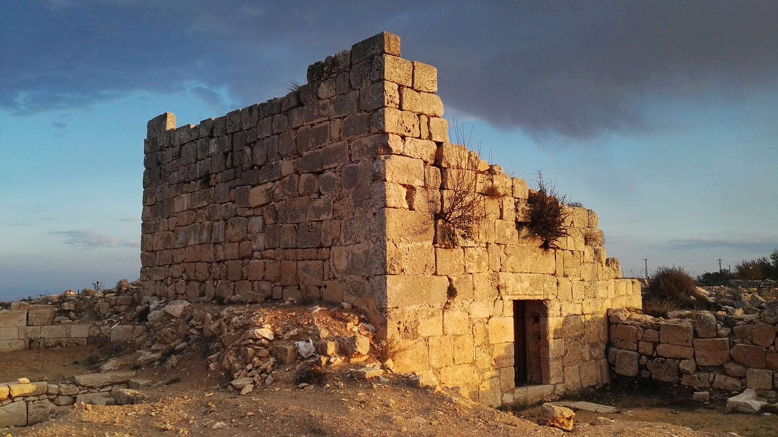 A ruined stone tower with a doorway at Burj Beitin, lit by low evening sun under a cloudy sky.