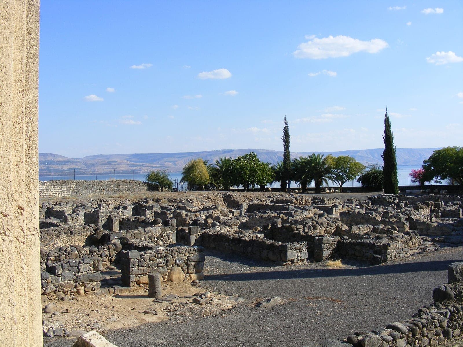 The low dark stone ruins of Capernaum, with the Sea of Galilee and hills behind.