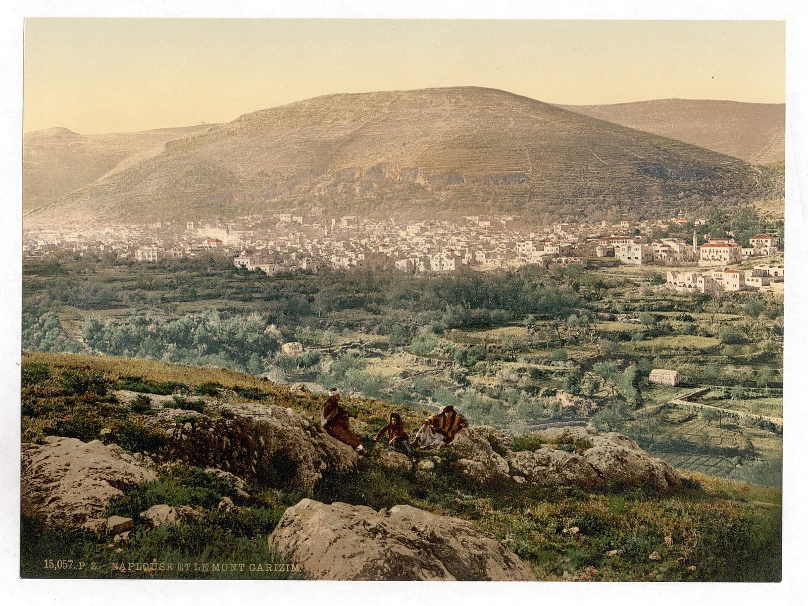 An old color photograph of Nablus in a green valley below Mount Gerizim, with people resting on rocks in front.