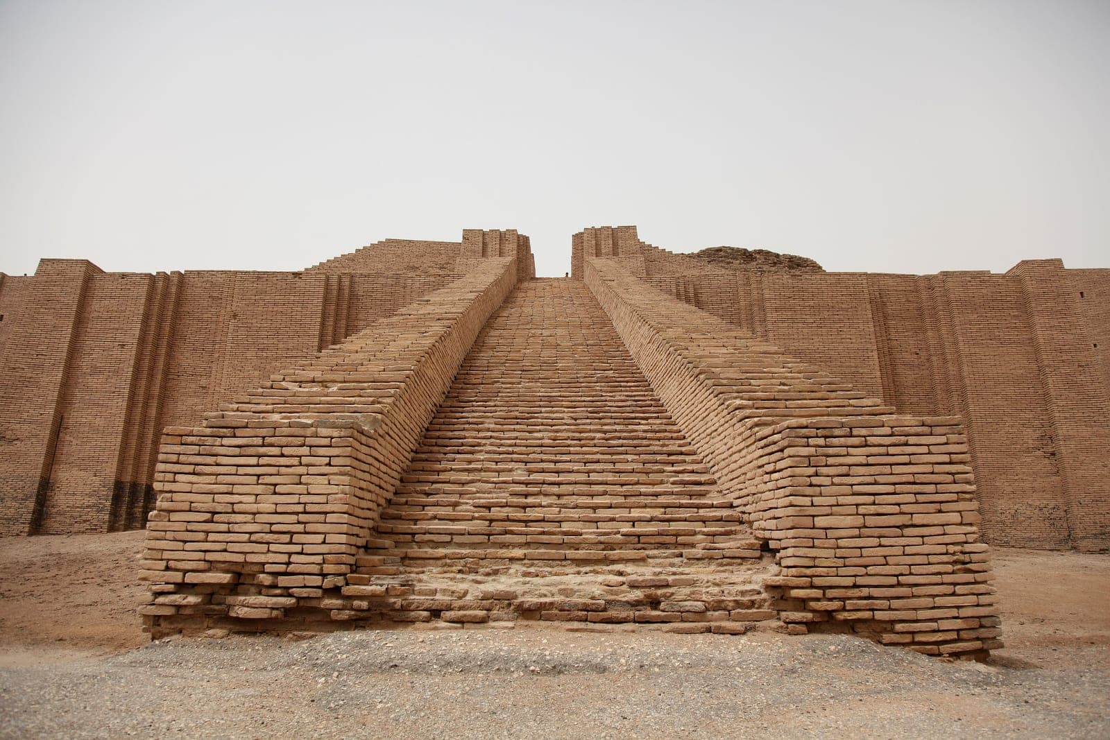 Wide frontal view of the Great Ziggurat of Ur, with a central brick stair rising between broad buttressed mud-brick walls in a hazy desert setting.
